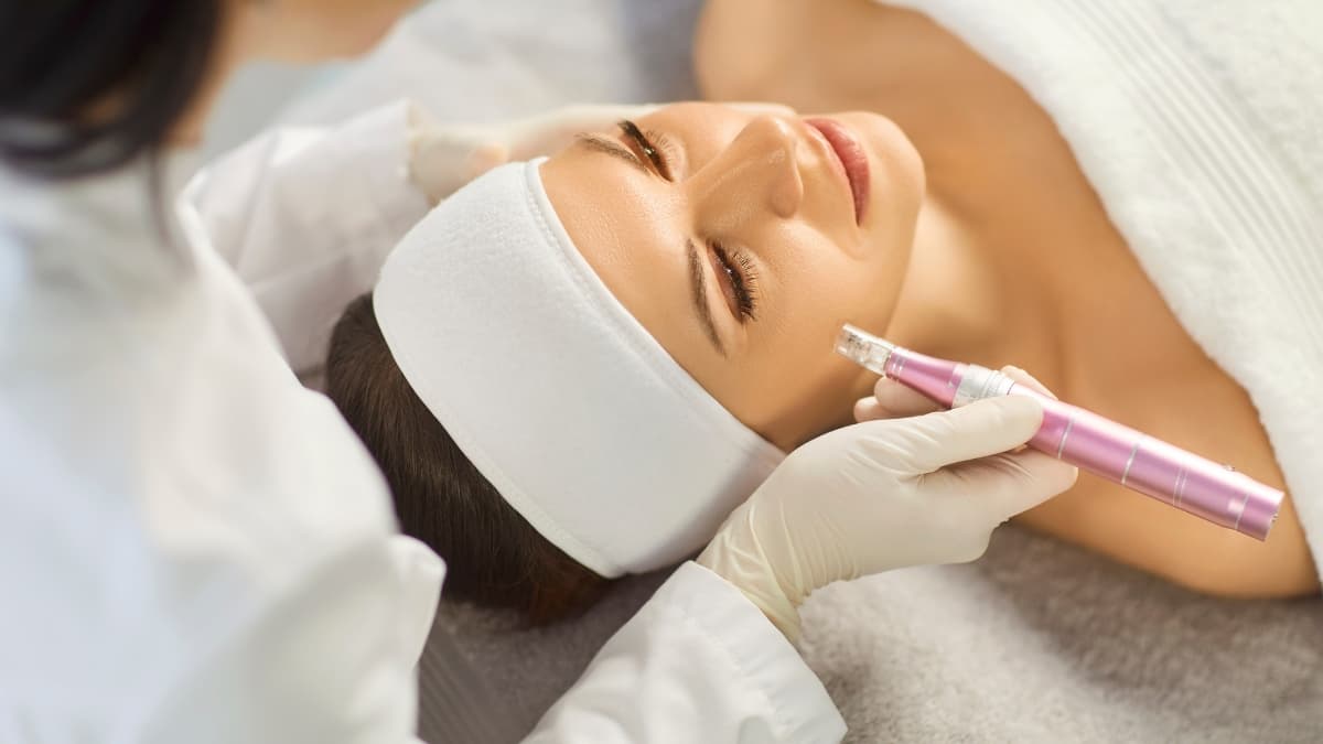 A woman receiving a professional microneedling treatment from an aesthetician using a pink dermapen while lying on a spa bed with a headband on.