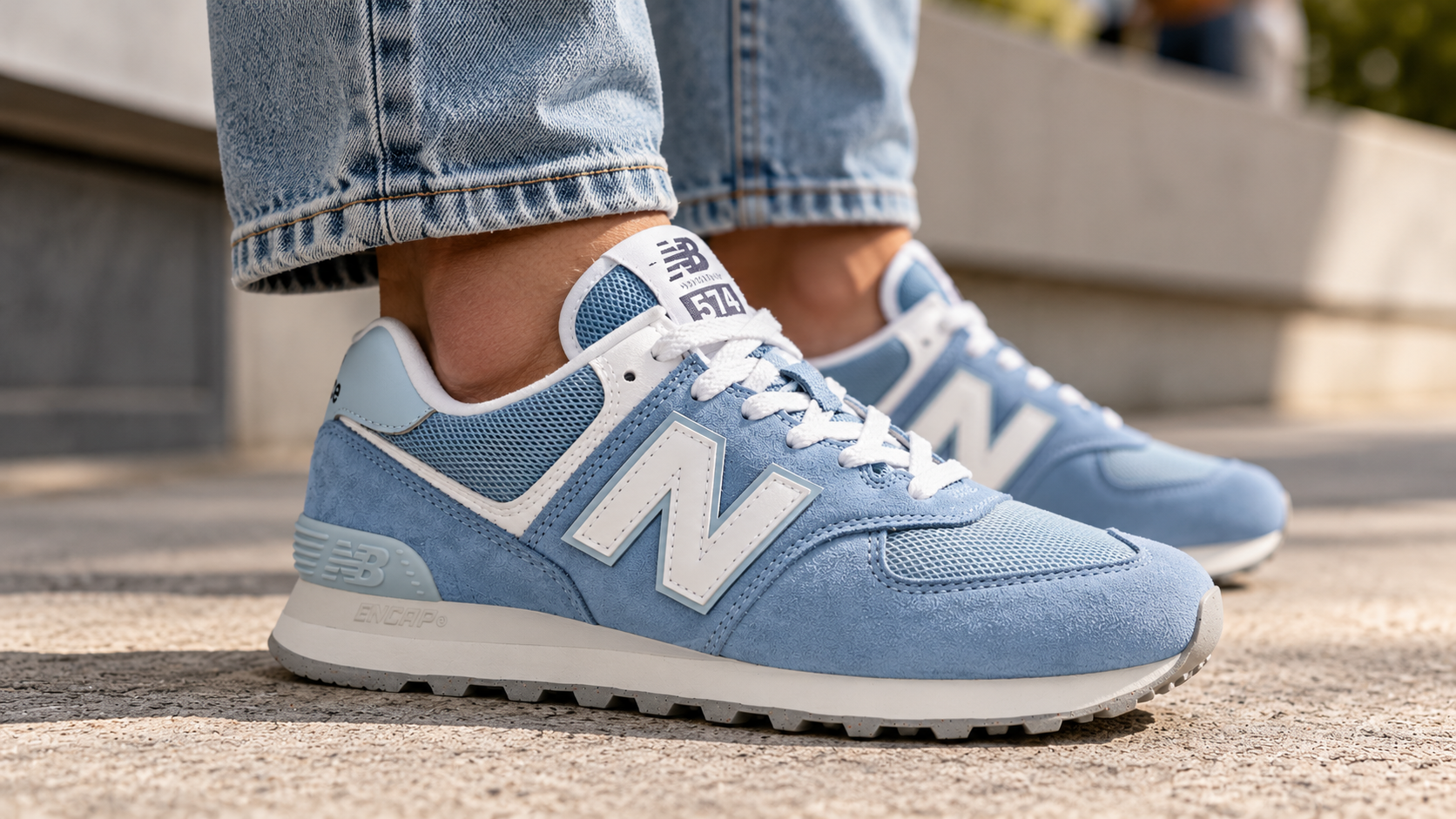 Person wearing light blue New Balance 574 sneakers with suede and mesh uppers, white laces, and ENCAP cushioning, styled with casual denim jeans on a sunlit urban sidewalk.